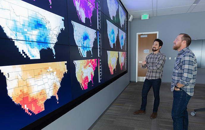 Two researchers stand next to a large screen displaying color-coded data on several U.S. maps