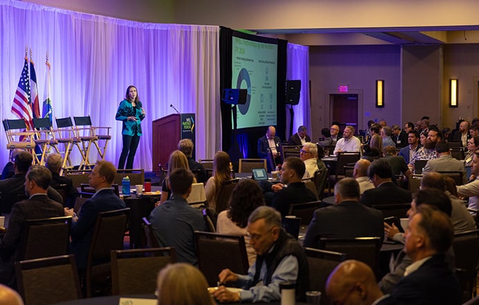 A person on a stage speaks to a room of people.