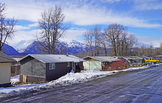 A row of houses surrounded by snow along a muddy road with mountains in the background