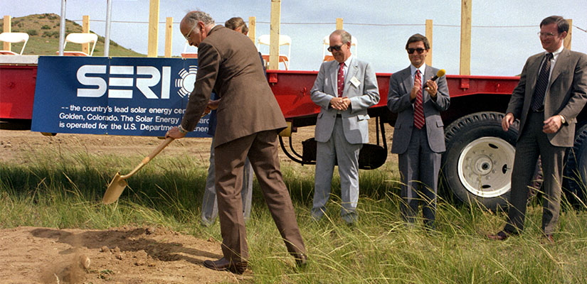 A person in a suit shovels dirt while others in the background applaud.