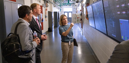 partnerships-93954 Two people look at a wall with monitors as another stands in front of them and gestures toward the screens.