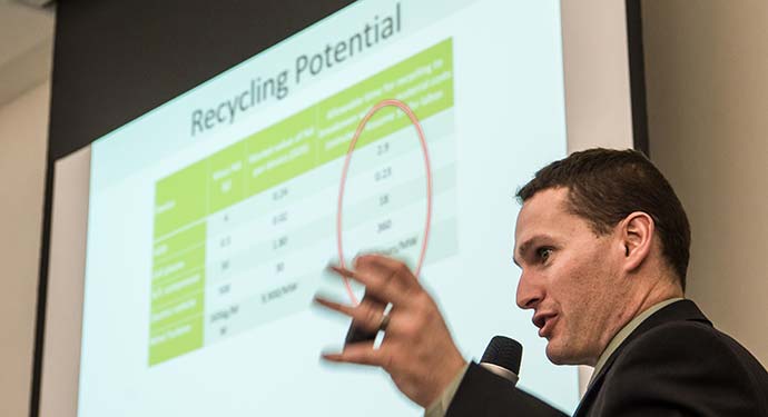A man speaks into a microphone in front of a slide presentation on recycling potential.