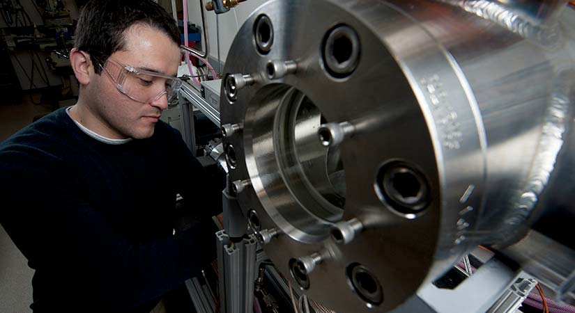 A researcher wearing protective eyewear works on a silver piece of machinery.