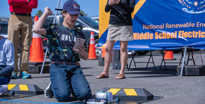 A child pumps afist in the air while holding a cellphone and kneeling near a track containing a miniature vehicle.