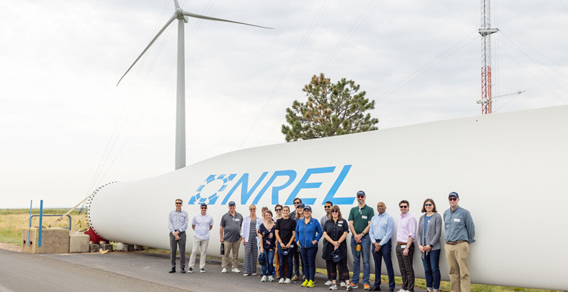 A group of people pose in front of a wind turbine and a large horizontal object labeled 'NREL'.