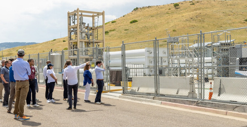 A person gestures to a fenced-in area containing various structures and equipment as a group looks on.