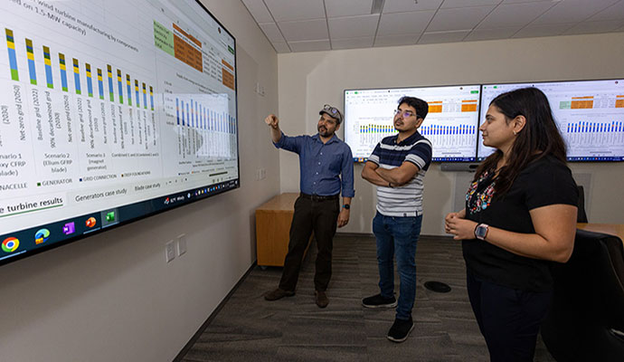 Three people stand and look at data on large display screen