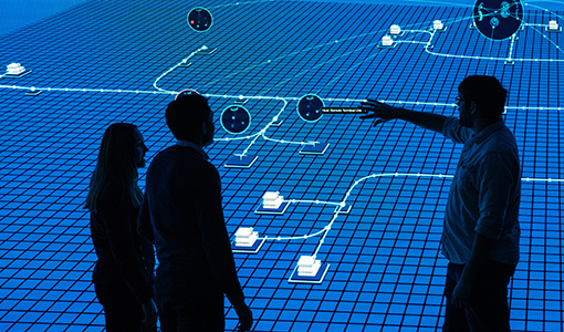 Three people stand in front of an point at large screen with grid data on display.