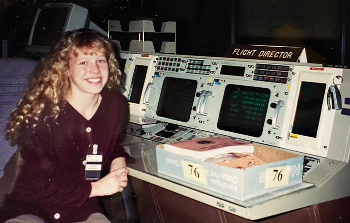 A high school girl sits at a space flight control training desk with a sign that says "Flight Director" 
