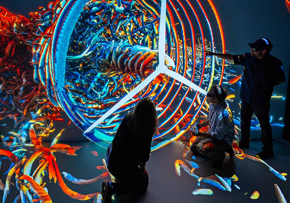 three researchers in silhouette interacting with a multicolor 3D visualization featuring a wind turbine.