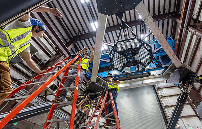 Four people wearing safety vests and hard hats work on ladders around a large piece of research equipment inside a laboratory building.