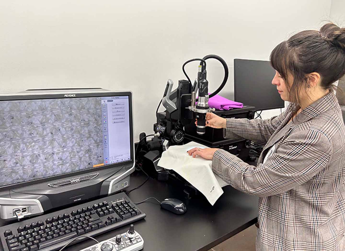 A person examines a fabric under a microscope next to a computer screen.