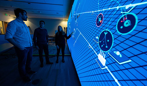 Three NREL cybersecurity researchers stand in front of a wall-sized projection demonstrating the Advanced Research on Integrated Energy Systems (ARIES).