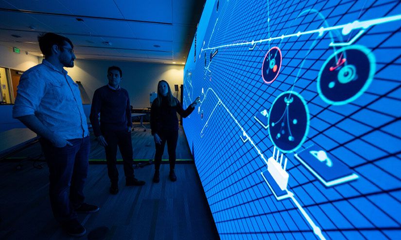 Three NREL cybersecurity researchers stand in front of a wall-sized projection demonstrating the Advanced Research on Integrated Energy Systems (ARIES).