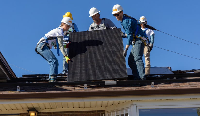 Five people in hard hats stand on a roof carrying a large square object.