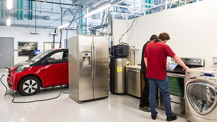 facilities-38895 Two men looking down at a laptop while standing near kitchen appliances and a charging electric vehicle.