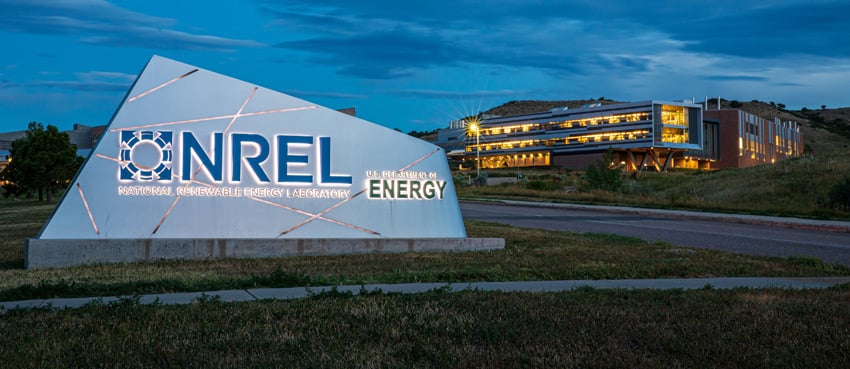 An entryway sign reading NREL - National Renewable Energy Laboratory - U.S. Department of Energy, with a lit-up building behind it.