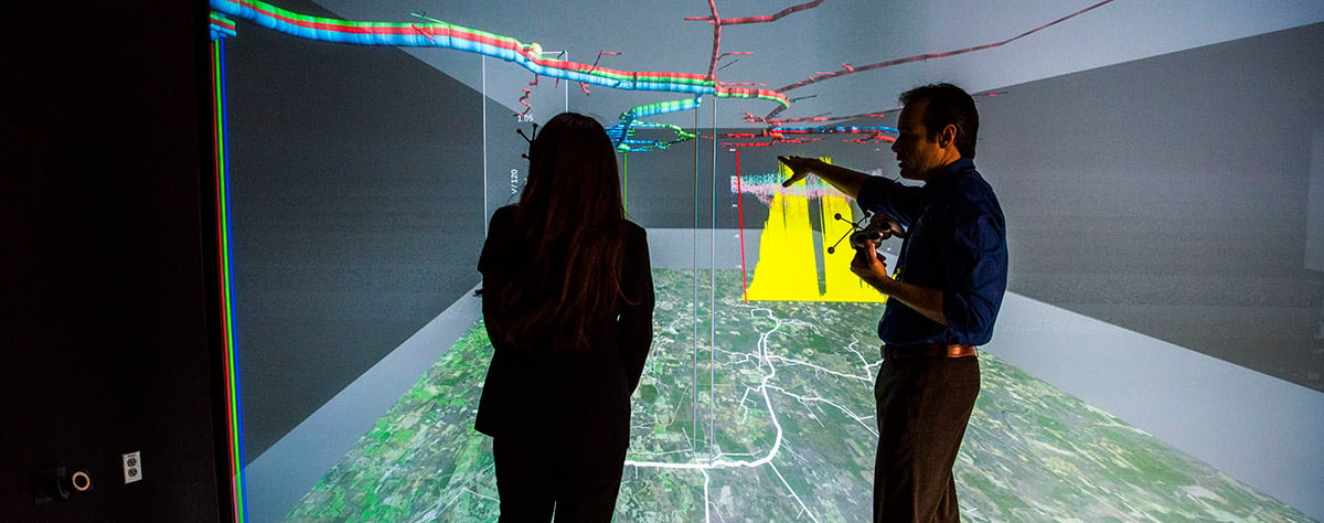 Two people standing in front of a large, color 3D visualization screen that spans the height and width of the wall they are facing.