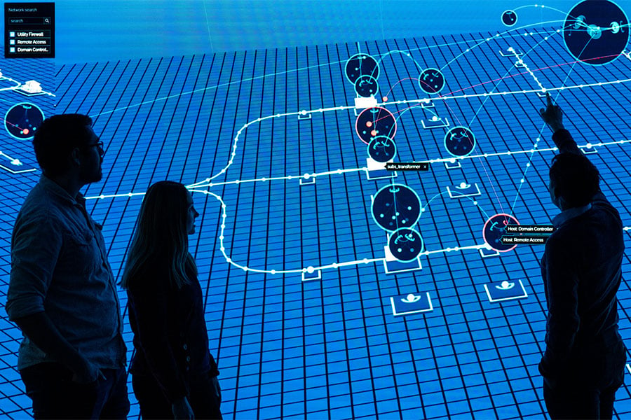 Three cybersecurity researchers stand in front of and point to digital display inside a research lab.