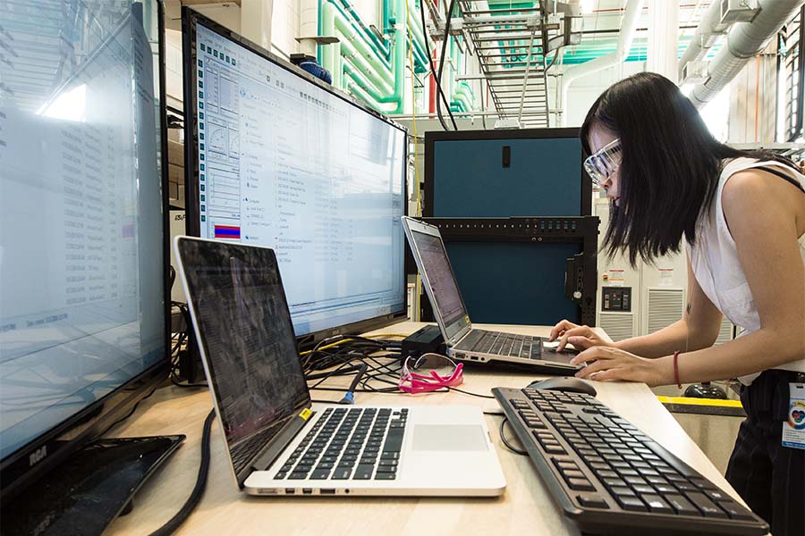 Person bends down to work on laptop on table with multiple computers and screens displaying data.