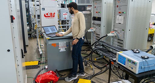 Person operates a laptop computer surrounded by power hardware, wires, and plugs in a laboratory.