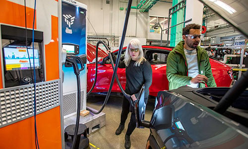 Two people charge electric vehicle inside laboratory and check under the hood.