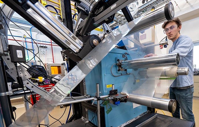 A researcher in a laboratory watching a continuous sheet of material run around multiple metal rolls.