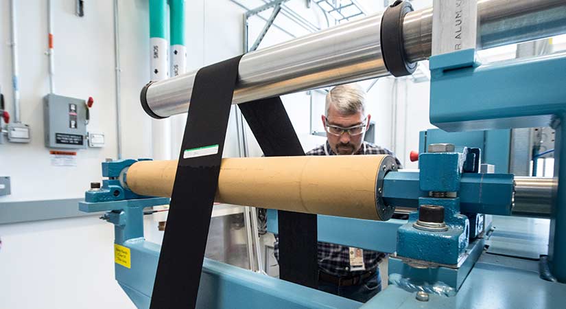 A photo of a man in a laboratory watching a black material run around various metal rolls.