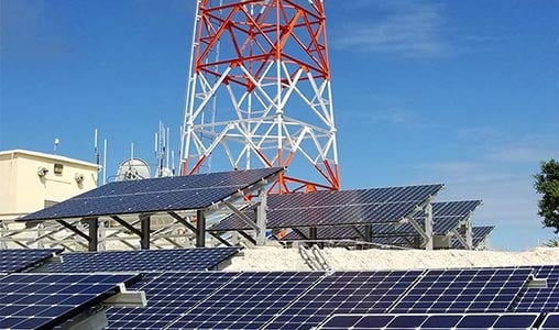 Rows of solar panels with a red and white tower on top of a building behind them.