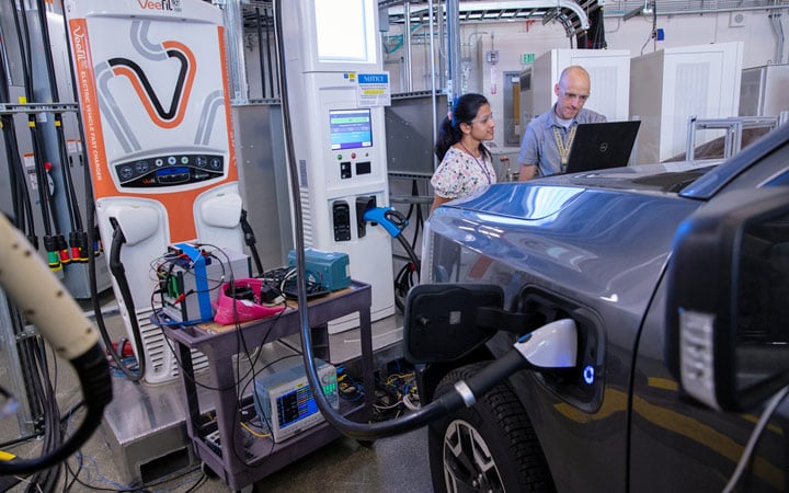 Two people wearing eye protection look at a laptop propped on the hood of an electric vehicle as it charges inside a laboratory.