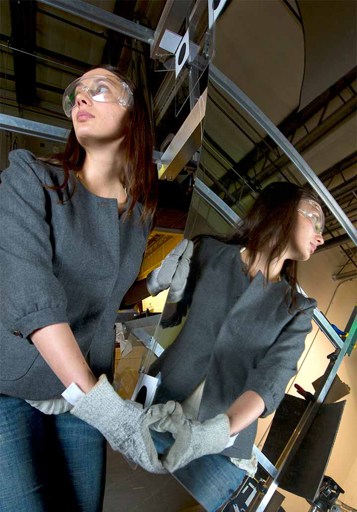 A woman in safety glasses is reflected in the large mirror she is moving with gloves.