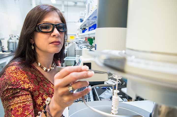 A woman in safety glasses reaches toward a piece of laboratoy equipment.