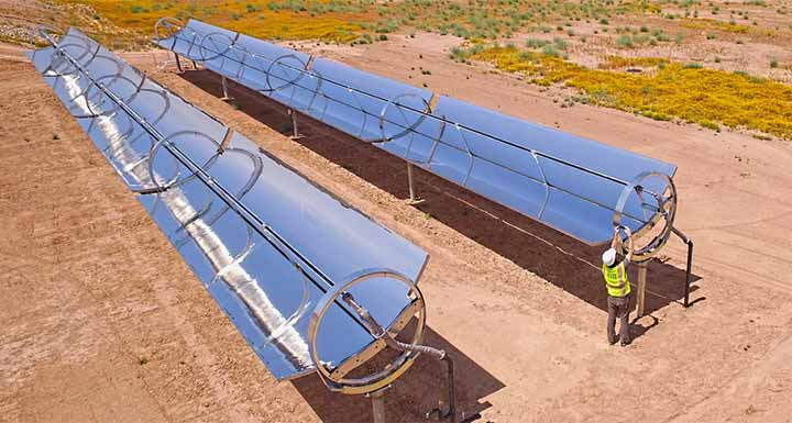 A man in a hardhat and reflective vest works on one of two reflective parabolic troughs amidst a desert landscape.