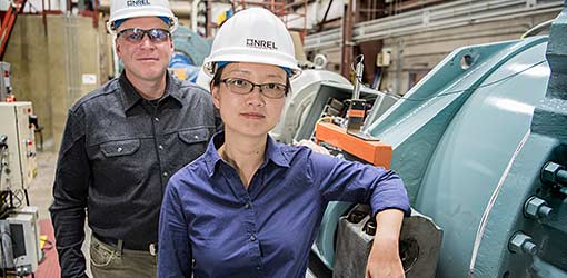 Photo of a man and a woman wearing hard hats and standing next to wind turbine testing equipment.