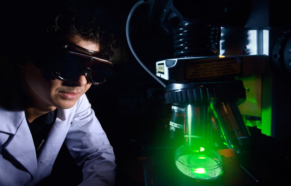 A person wearing eye protection examines liquid in a glass beaker.