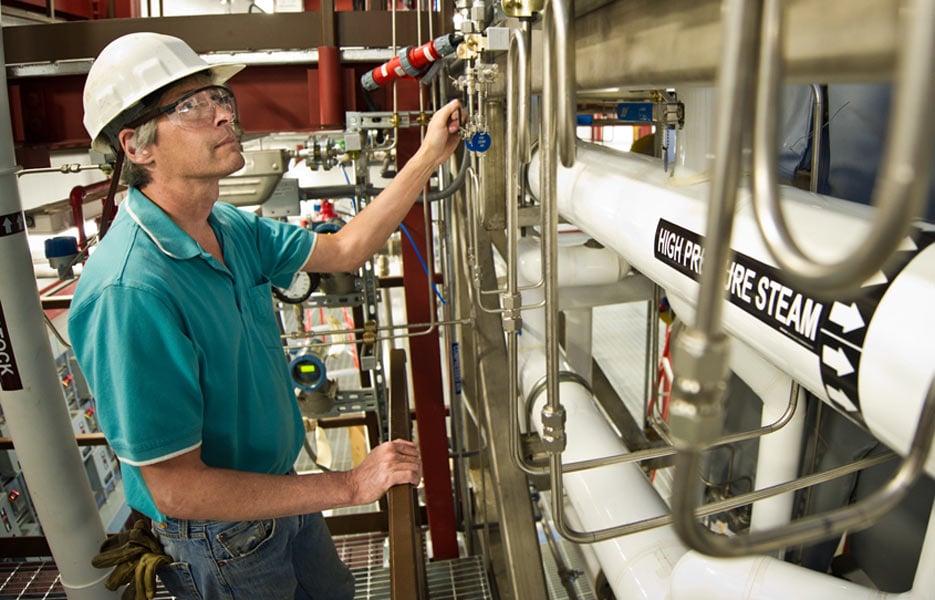 A person in a hardhat and eye protection stands among laboratory equipment.