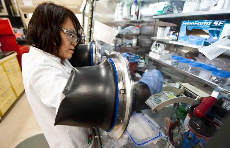 A person in a lab coat wearing eye protection works within a laboratory.