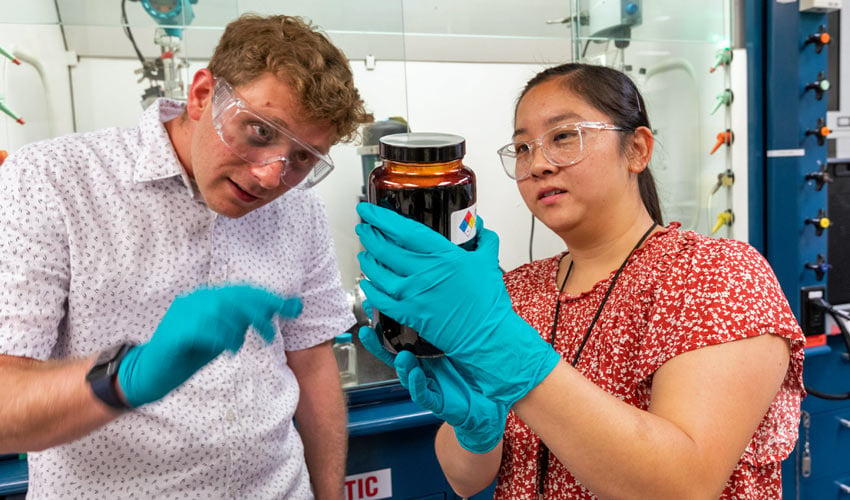 One person wearing gloves holds a jar containing dark material while they and another person look at its contents.