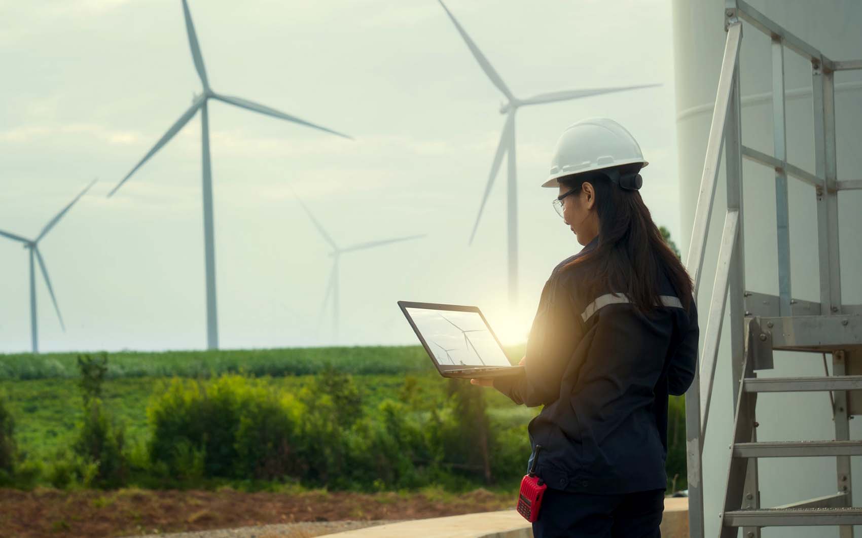 : A person dressed in a construction outfit looks at a tablet with a picture of wind turbines on the screen. In the distance, there is a field with four staggered wind turbines.