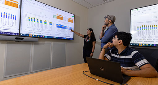 Three people looking at laptop and large data display screen