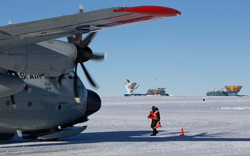 A person places orange safety cones on a snowy runway next to a large cargo plane with large telescopes in the background.
