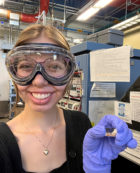 A person smiling in a laboratory holding a photovoltaic device with a gloved hand. 