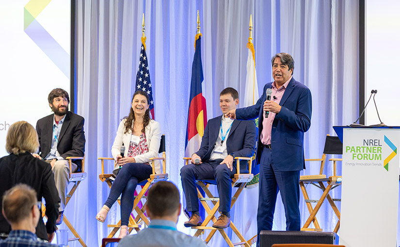 Three people sit on a stage smiling and laughing while a moderator points to them with people watching from the crowd.