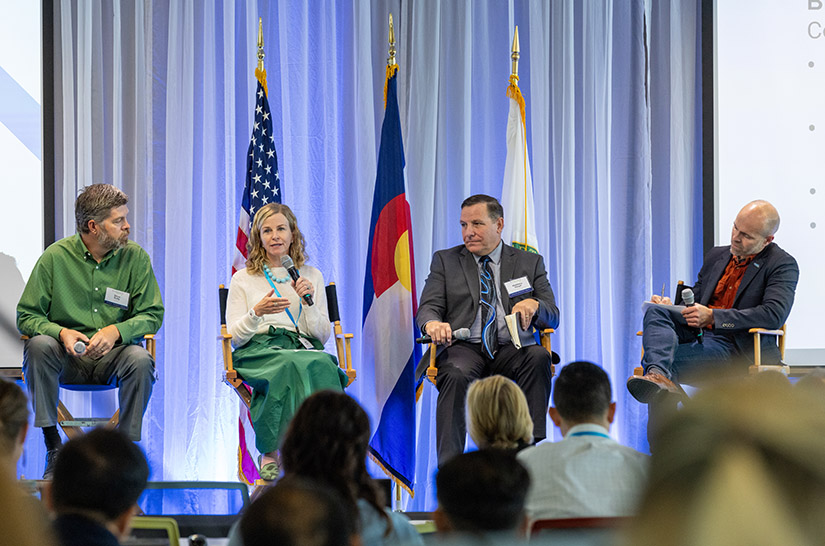 A woman seated on a stage address a crowd while three men seated on the stage listen and take notes.