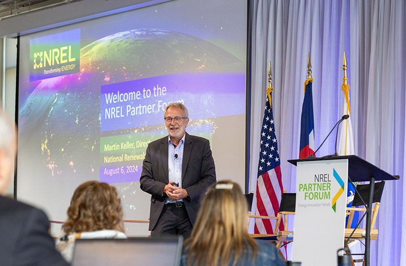 A man with a white beard and glasses addresses the crowd in front of a screen welcoming them to the Partner Forum. 