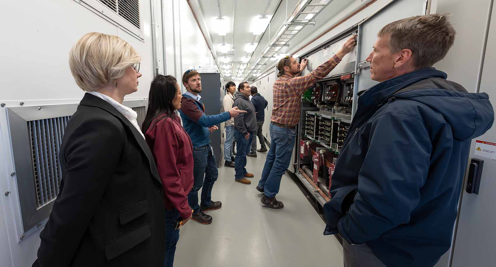 Several people in a hallway with a wall of electrical equipment.