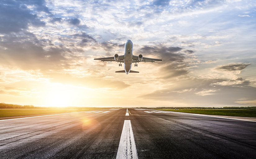 A plane flies over a runway in front of a sunrise.