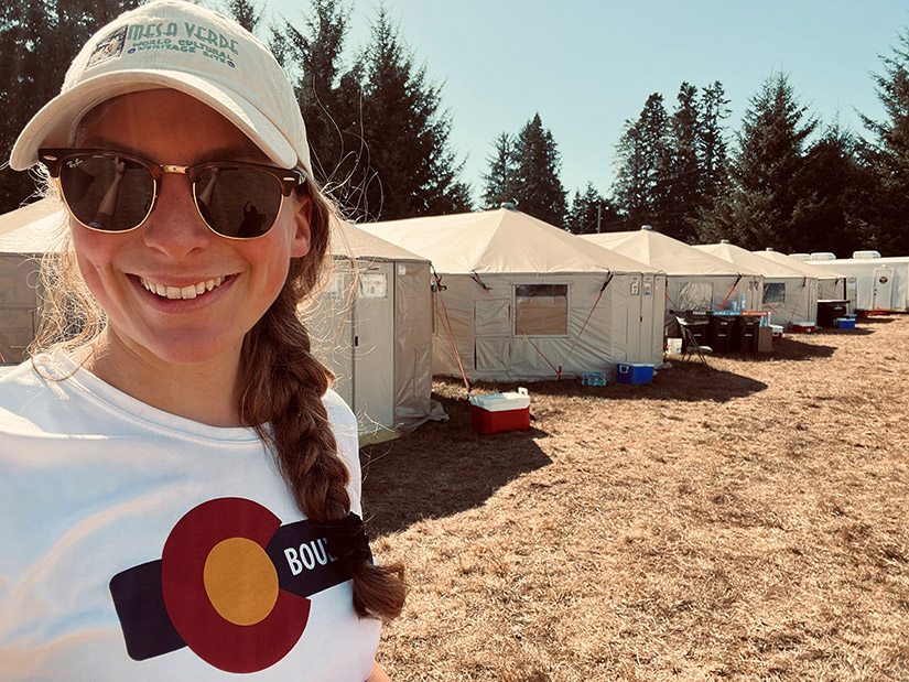 A smiling person wearing sunglasses poses for a selfie in front of a row of fire camp yurts.