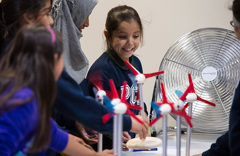 A group of students look at model wind turbines with a fan behind them. 