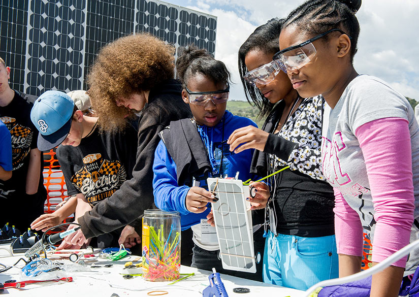 Several high school students standing at a table outside build miniature solar-powered race cars.